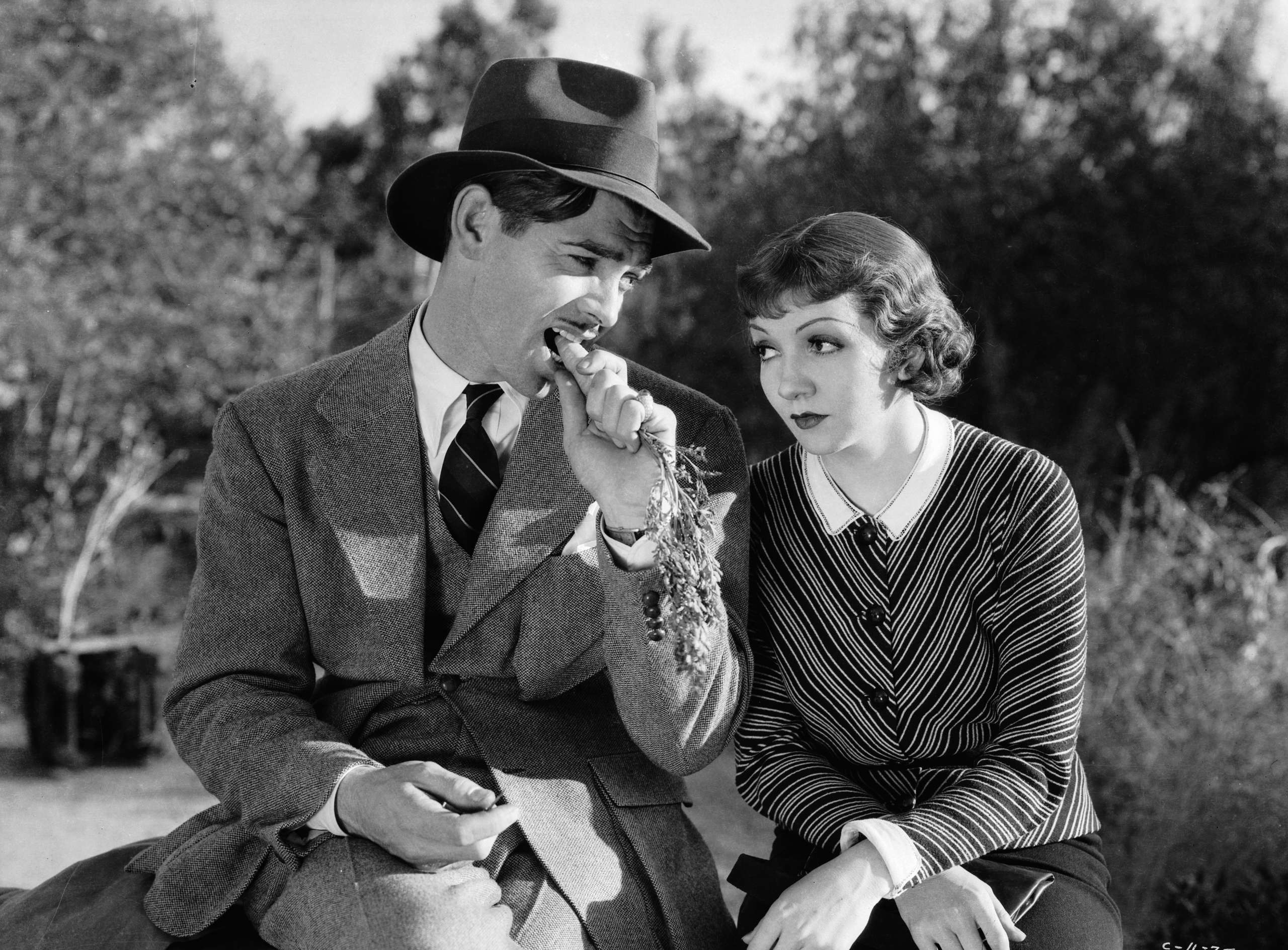 1934:  Socialite Ellie Andrews, played by Claudette Colbert (1903 - 1996) appears distracted while reporter Peter Warne, played by Clark Gable (1901 - 1960), bites nonchalantly on a carrot in Frank Capra's romantic comedy, 'It Happened One Night'.  (Photo via John Kobal Foundation/Getty Images)